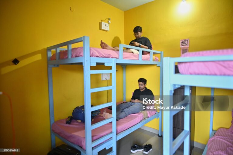 A picture of travelers staying in a hostel. They are in a room with bunk beds, likely sharing the space. They are relaxing and using their mobile phones after a tiring day. The room has a casual and friendly atmosphere.